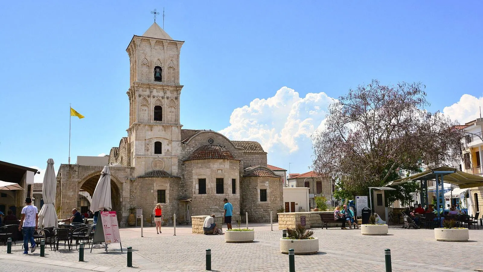 The stone bell tower and domes of the Church of Saint Lazarus in Larnaca, Cyprus, on a sunny square with cafe tables and a flowering tree.