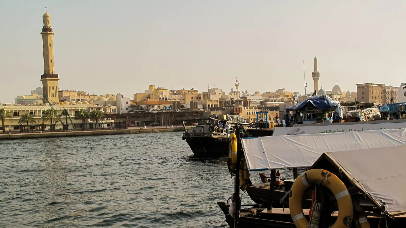 Wooden abra ferry boats moored on Dubai Creek with the minaret of the Grand Mosque and the low sand-coloured buildings of old Bur Dubai behind them.