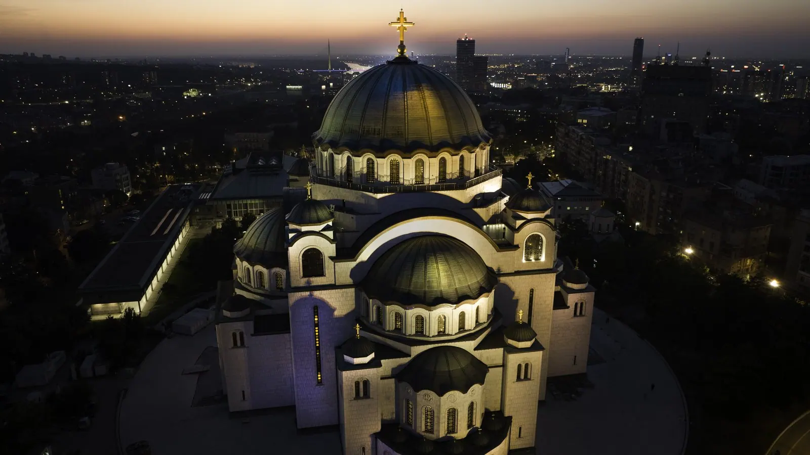 Biserica Sfântul Sava din Belgrad, Serbia, văzută de sus la amurg, cu cupolele albe luminate și crucea aurită deasupra luminilor orașului.