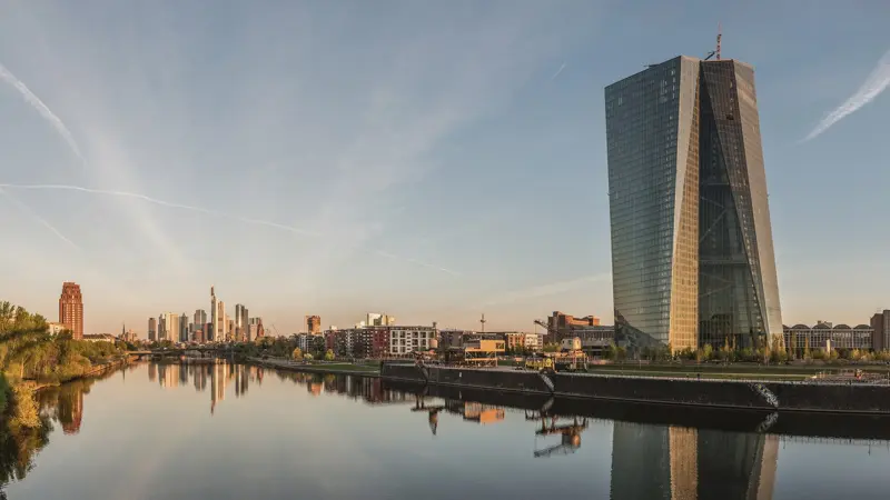 Turnul de sticlă al Băncii Centrale Europene pe malul râului Main, la Frankfurt, în zori, cu silueta orașului și reflexia ei în apă.
