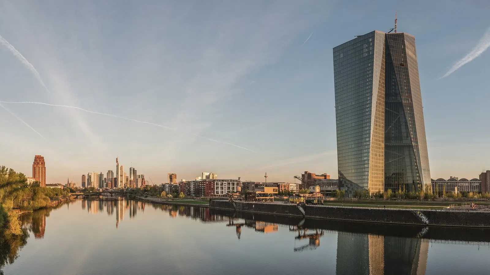 The glass tower of the European Central Bank beside the river Main in Frankfurt at dawn, with the city skyline and its reflection in the water.