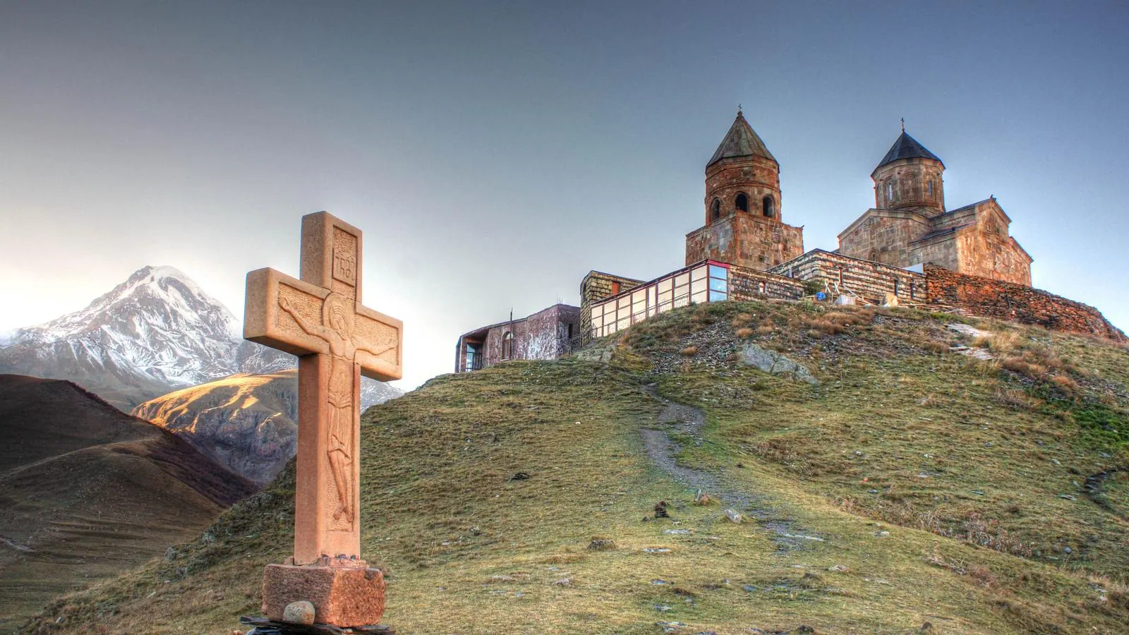 Biserica Sfânta Treime din Gergeti pe o culme verde, sub vârful înzăpezit Kazbek din Georgia, cu o cruce de piatră sculptată în prim-plan.