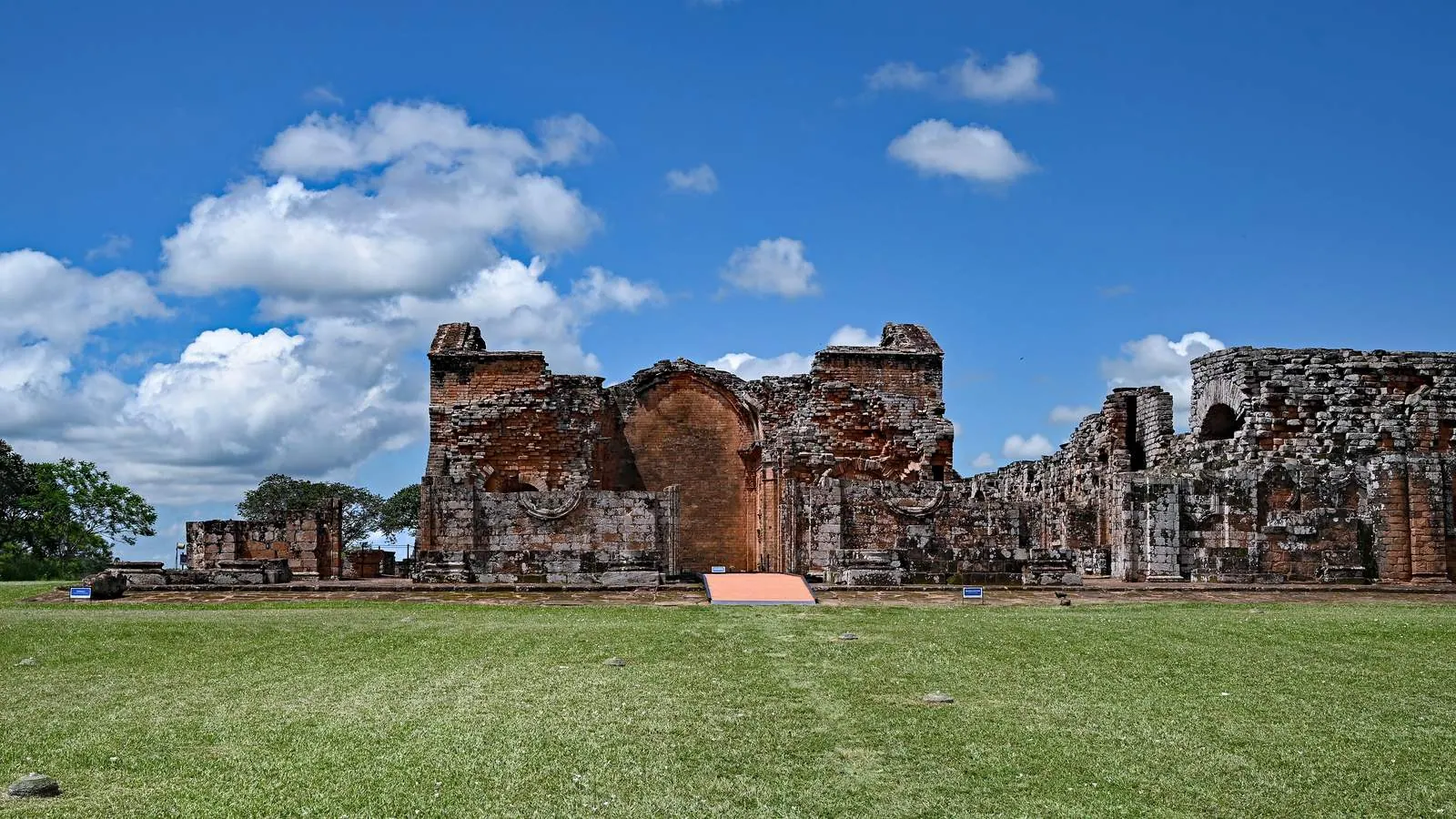 Ruinele din gresie roșie ale bisericii misiunii iezuite La Santísima Trinidad de Paraná din Paraguay, sub un cer albastru cu nori albi.