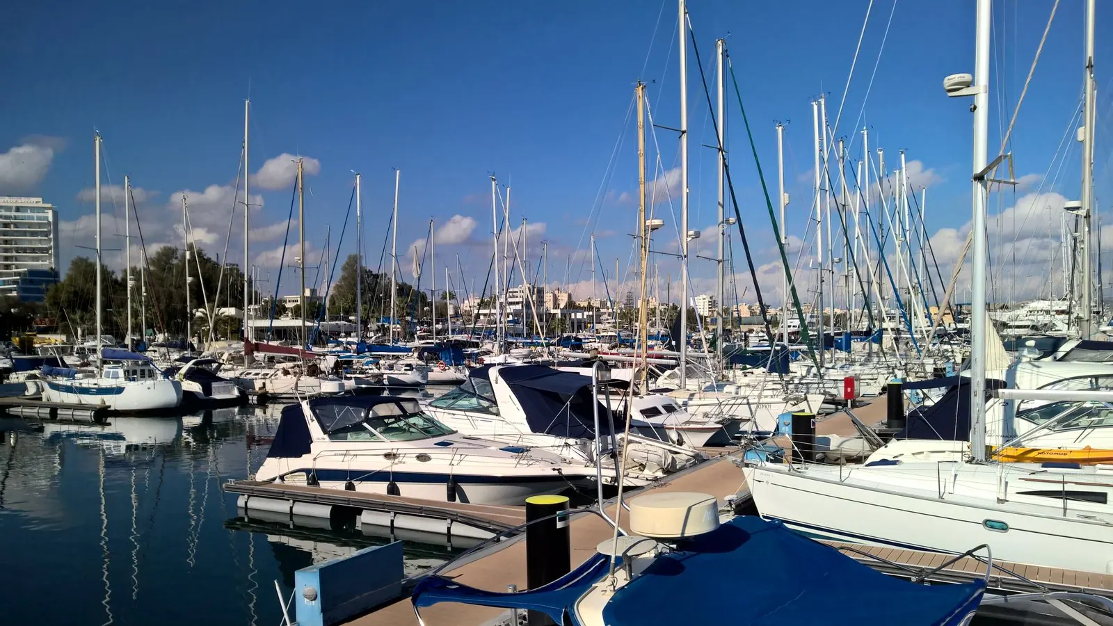 Sailing boats and motor yachts moored at Larnaca Marina, Cyprus, on a sunny day, with the seafront buildings behind under a blue sky.